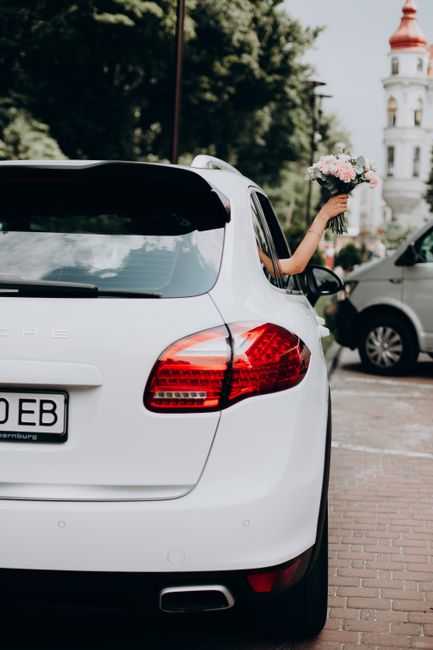 A person is extending their arm out of the driver’s side window of a white Porsche car, holding a bouquet of fresh flowers. The bouquet features soft pink roses, white blossoms, and green foliage, w